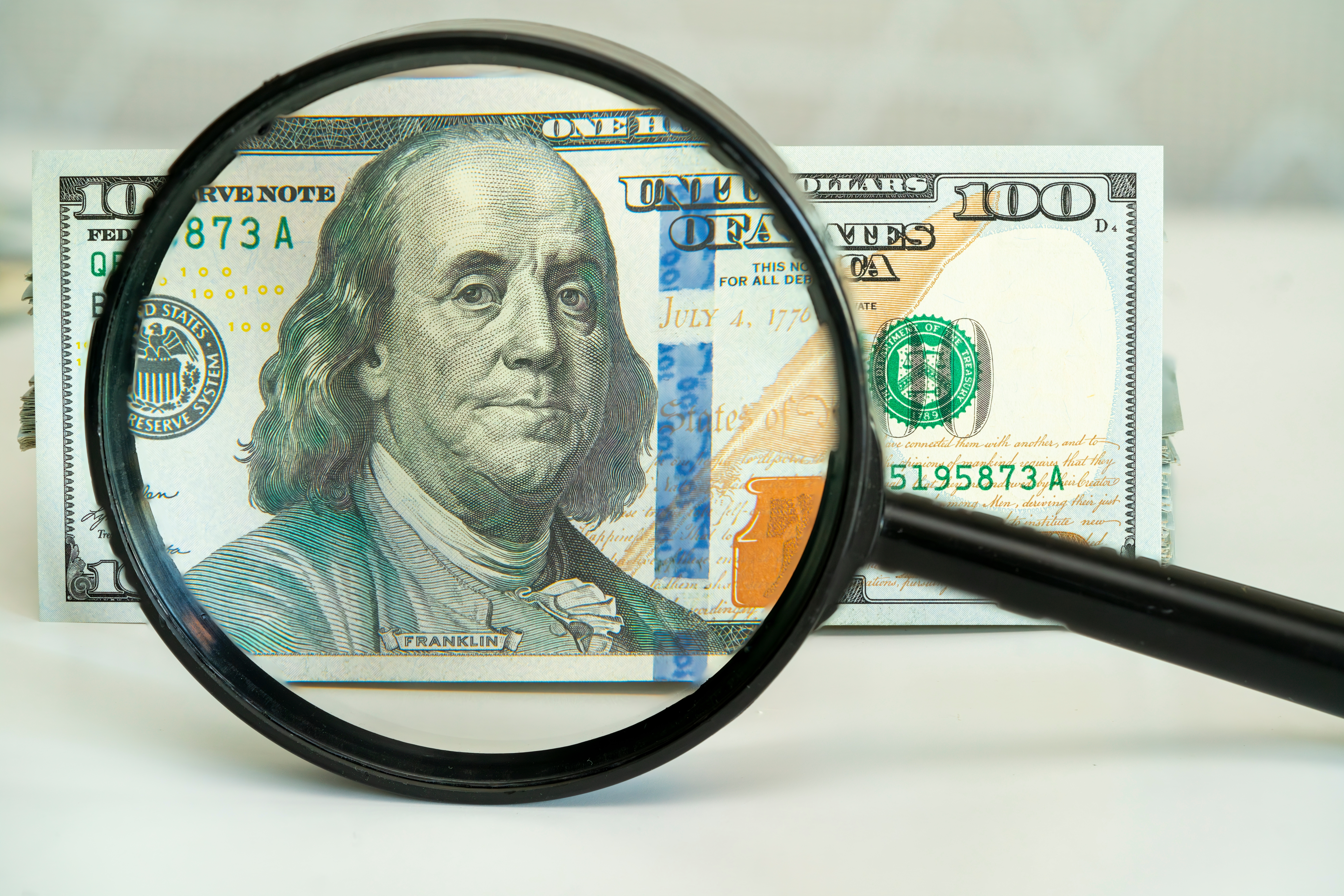 A magnifying glass hovers over a hundred dollar bill, revealing intricate details of the design, emphasizing Benjamin Franklin's portrait and security features.